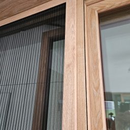 ROLETY -MaAn DOMINIKA SCHÜTZ - Złożona, plisowana siatka okienna w drewnianej ramie, widoczna faktura drewna, zbliżenie na detale konstrukcyjne.