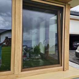 ROLETY -MaAn DOMINIKA SCHÜTZ - Drewniane okno z zamontowaną siatką przeciw owadom, widok na ogród i odbicie fotografa w szybie.