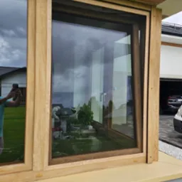 Drewniane okno z zamontowaną siatką przeciw owadom, widok na ogród i odbicie fotografa w szybie.