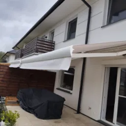 Biała, rozłożona osłona przeciwsłoneczna zamontowana nad tarasem nowoczesnego domu z widokiem na ogród z basenem.