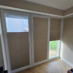 Beżowe, plisowane osłony okienne zamontowane na trzech oknach w jasnym pomieszczeniu z parkietem ułożonym w jodełkę.