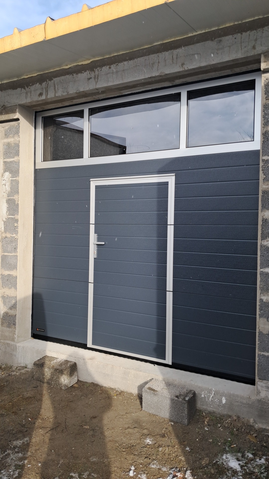 Ciemnoszara segmentowa brama garażowa z drzwiami wejściowymi, zamontowana w surowym, betonowym otworze z oknami nad bramą.