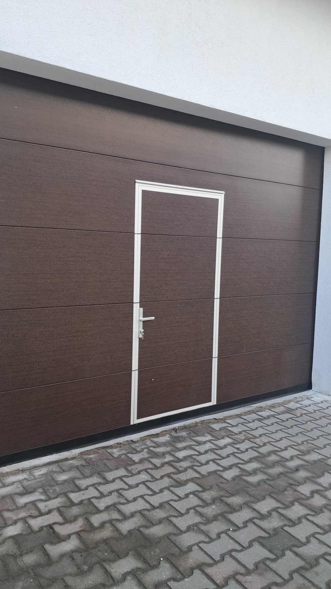 Ciemnobrązowa segmentowa brama garażowa z wbudowanymi drzwiami wejściowymi z białym obramowaniem i srebrną klamką na tle kostki brukowej.