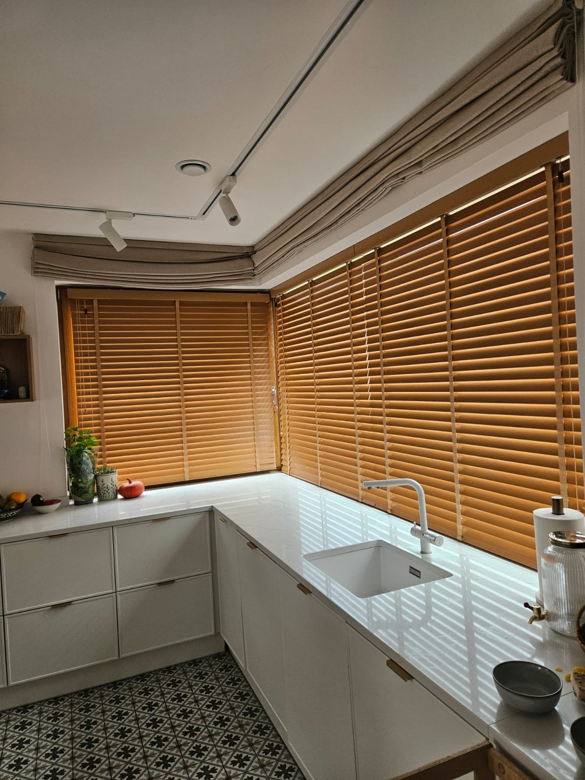 Drewniane żaluzje poziome zamontowane w oknach kuchni z białym blatem i zlewozmywakiem, widoczne także lampy sufitowe i zasłony rzymskie nad oknami.