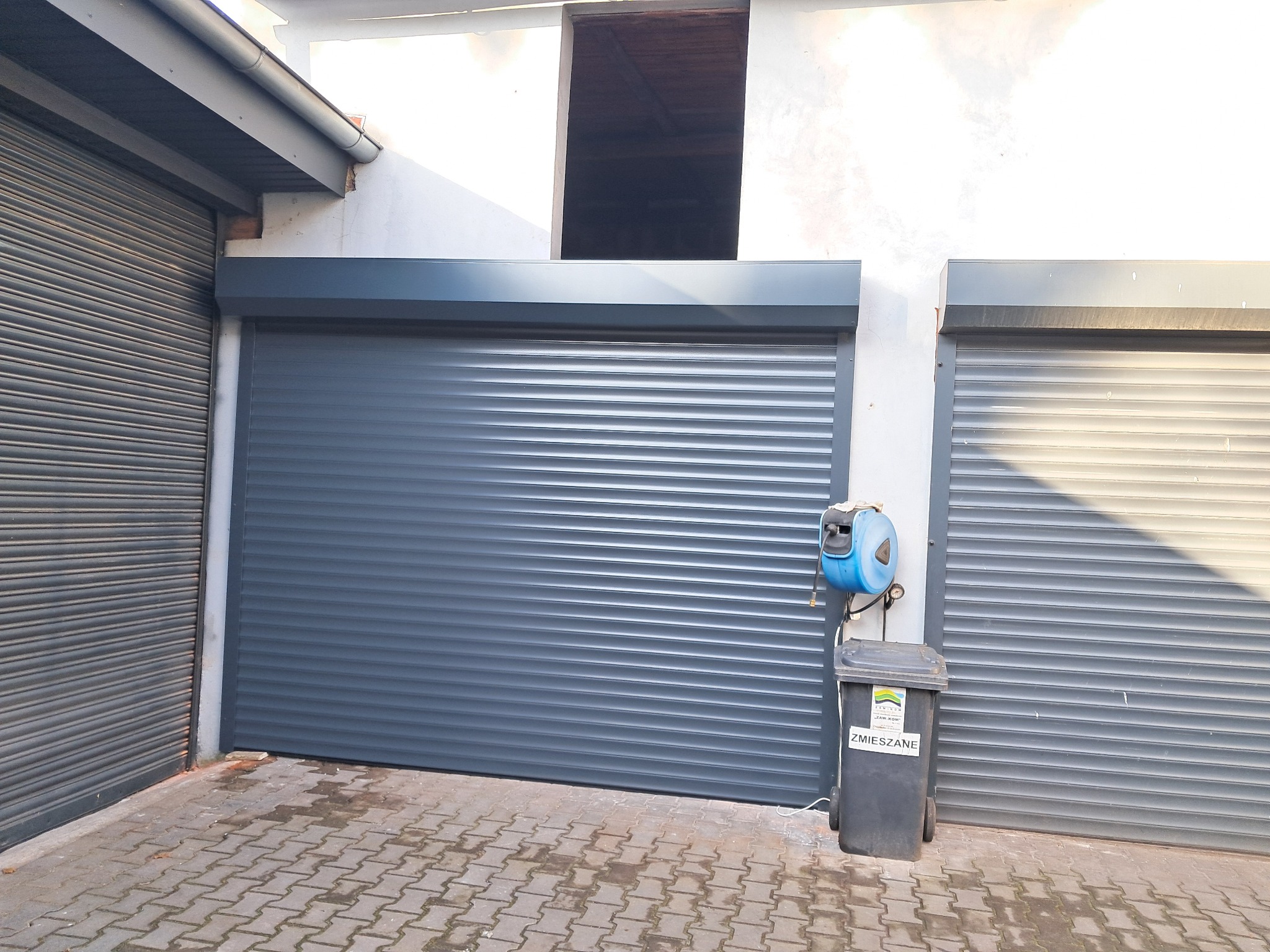 Szara, pozioma, zwinięta osłona na bramie garażowej, obok pojemnik na śmieci z napisem 'ZMIESZANE' i zwinięty wąż ogrodowy na ścianie.