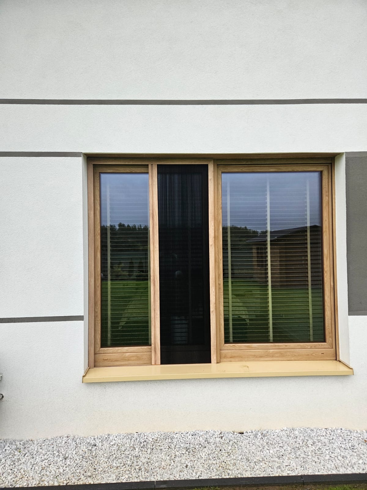 Okno z drewnianą ramą, wyposażone w moskitierę plisowaną, chroniącą przed owadami, z widokiem na zielony trawnik i drzewa w tle.