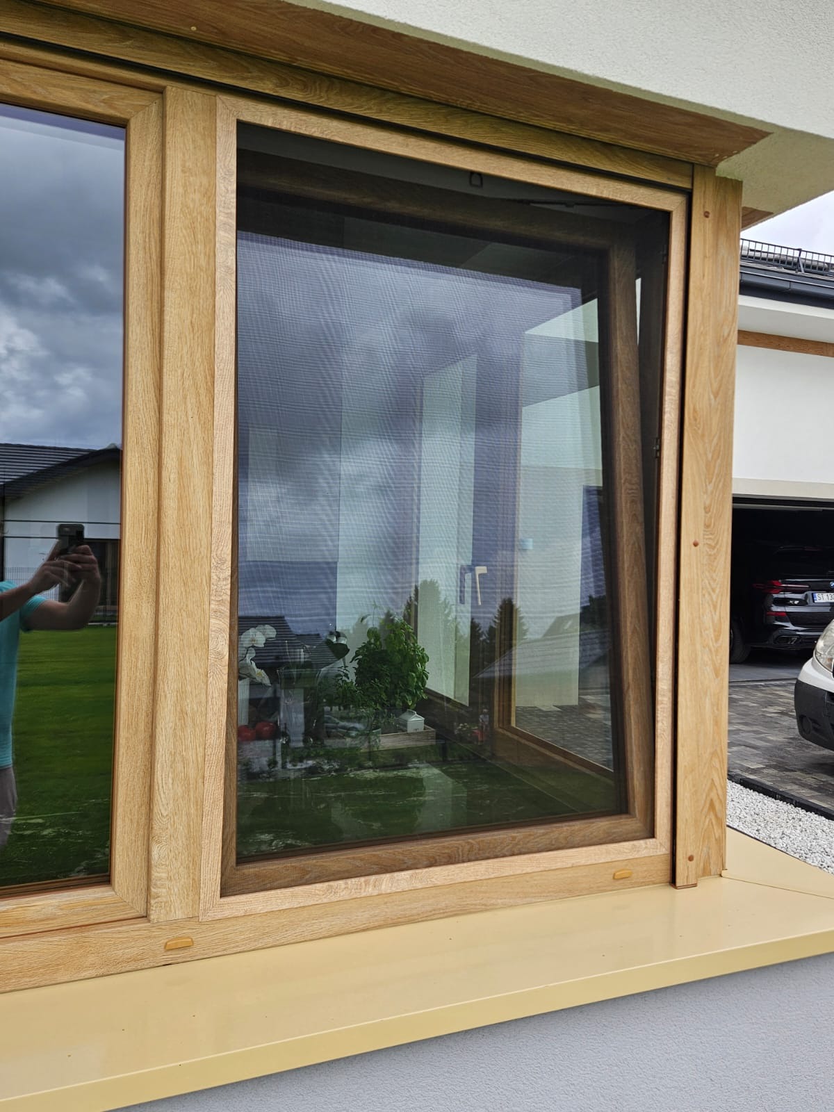 Drewniane okno z zamontowaną siatką przeciw owadom, widok na ogród i odbicie fotografa w szybie.