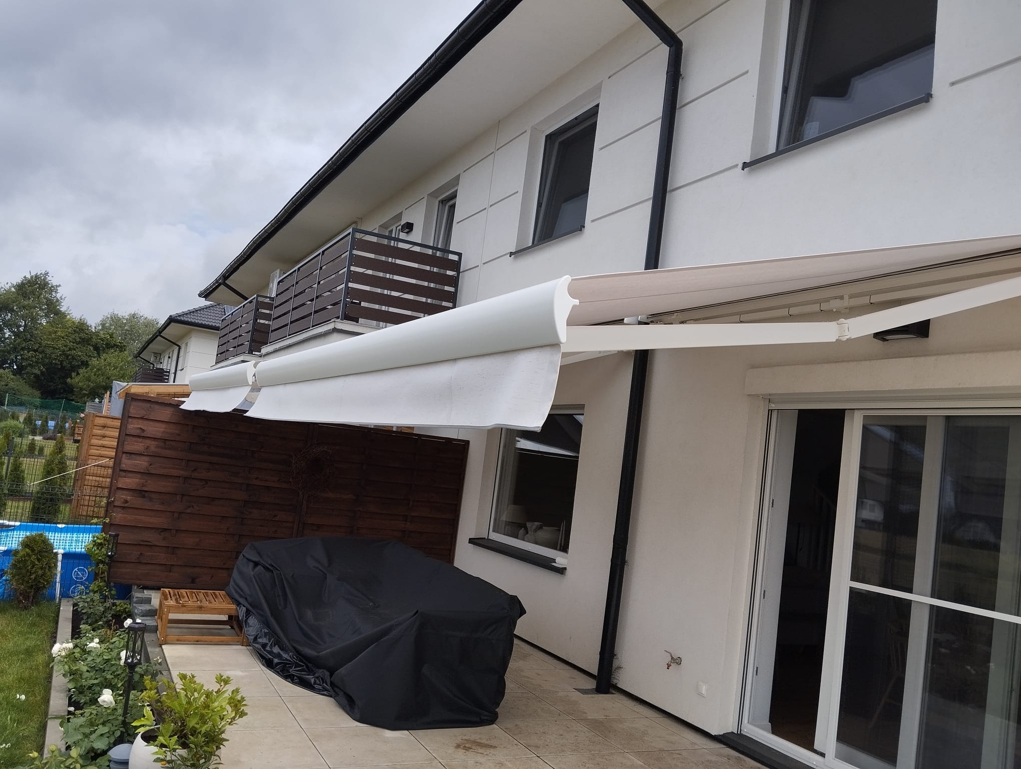 Biała, rozłożona osłona przeciwsłoneczna zamontowana nad tarasem nowoczesnego domu z widokiem na ogród z basenem.