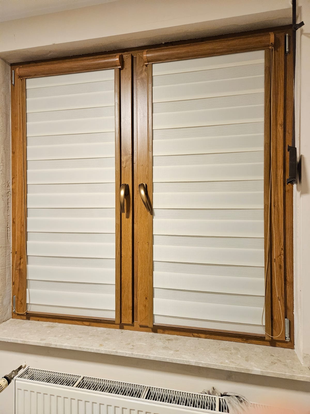 Zamknięte dwuskrzydłowe okno z drewnianą ramą i kremowymi poziomymi przesłonami w stylu dzień i noc, widoczny fragment grzejnika pod parapetem.