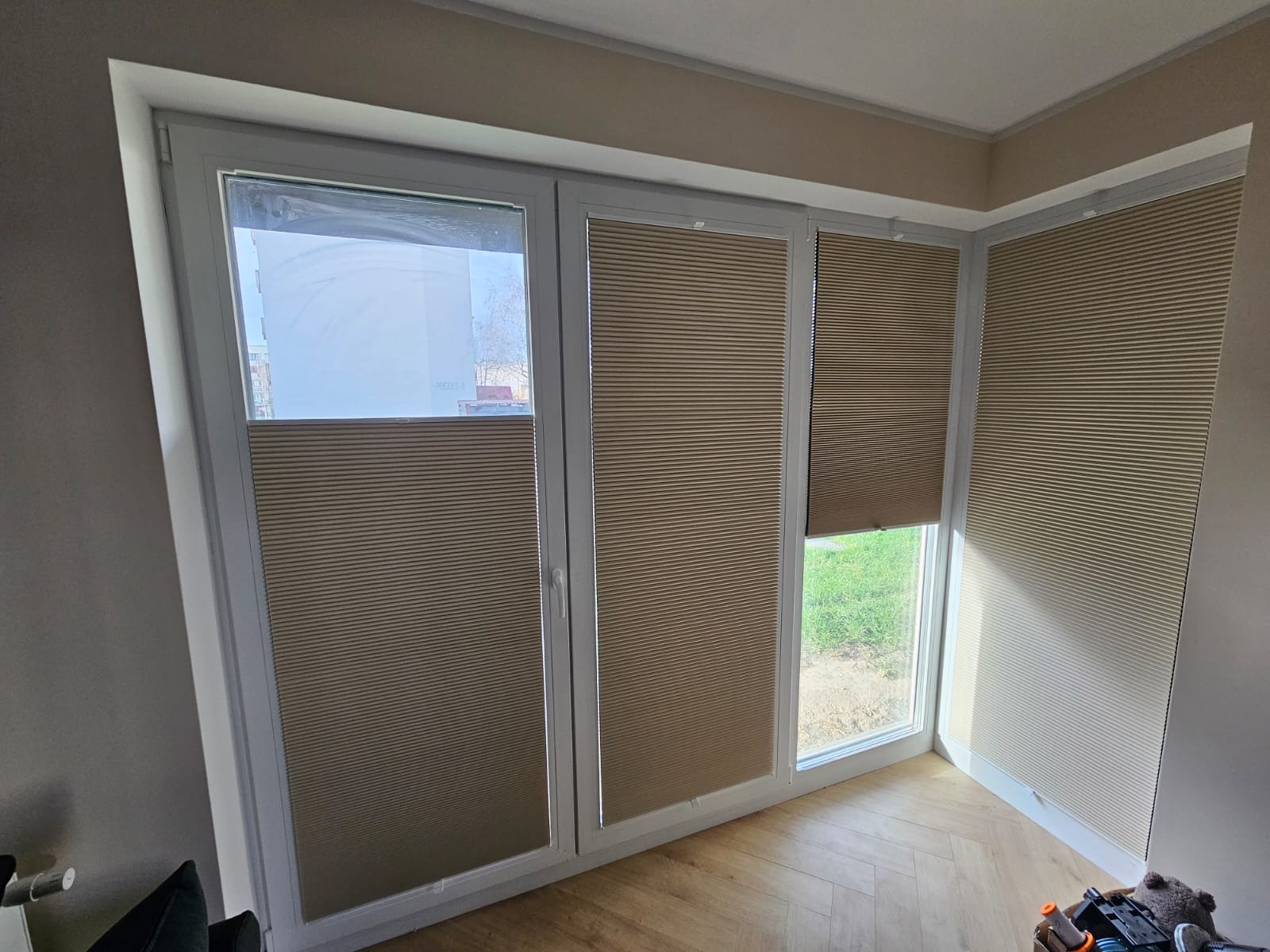 Beżowe, plisowane osłony okienne zamontowane na trzech oknach w jasnym pomieszczeniu z parkietem ułożonym w jodełkę.