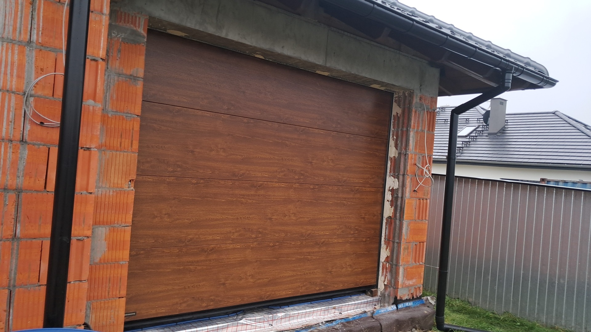 Montaż segmentowej bramy garażowej w surowym stanie, widoczne niedokończone mury z cegły i instalacja odprowadzania wody deszczowej.