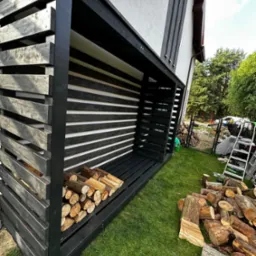 Czarna, drewniana konstrukcja na drewno opałowe, częściowo wypełniona, z widocznym stosem drewna obok, na tle trawnika i domu.