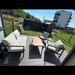 Pomorskie Tarasy - Nowoczesny taras z szarych desek kompozytowych. Zestaw mebli wypoczynkowych z jasnymi poduchami i stolikiem. W tle zielony trawnik i budynek mieszkalny.