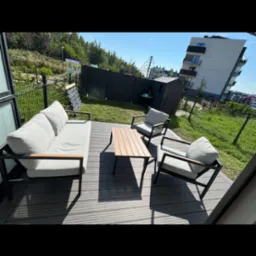 Nowoczesny taras z szarych desek kompozytowych. Zestaw mebli wypoczynkowych z jasnymi poduchami i stolikiem. W tle zielony trawnik i budynek mieszkalny.