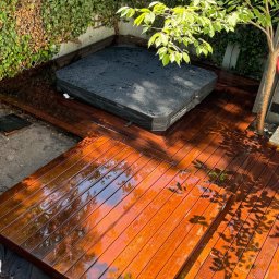 Pomorskie Tarasy - Błyszczący, drewniany taras z jacuzzi w ogrodzie, otoczony zielenią. Widoczne detale desek i odbicia światła. Ujęcie z góry ukazuje całą konstrukcję.