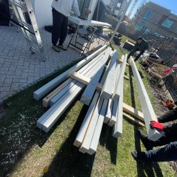 Pomorskie Tarasy - Przygotowanie elementów konstrukcyjnych na trawie do montażu, prawdopodobnie pergoli lub altany, z pracownikami w tle i drabiną obok.