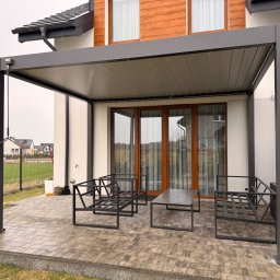 Pomorskie Tarasy - Nowoczesna, szara pergola z metalu przylegająca do domu. Na tarasie minimalistyczne meble ogrodowe i donica. Widok na osiedle domów jednorodzinnych w tle.