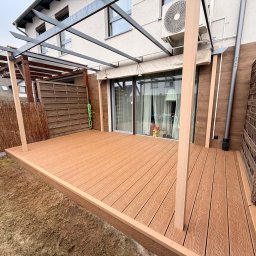 Pomorskie Tarasy - Jasny taras kompozytowy z metalową konstrukcją pergoli, widok z poziomu gruntu. Deski ułożone równolegle do ściany budynku, brązowe panele ogrodzeniowe po bokach.