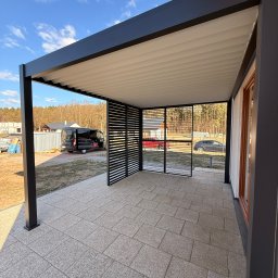 TARASY PÓŁNOCY - Nowoczesna, czarna pergola z lamelami i ażurowymi ścianami, osłaniająca taras z jasnej kostki. W tle dom i samochody, błękitne niebo.