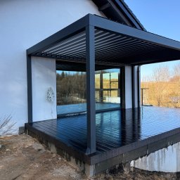 TARASY PÓŁNOCY - Nowoczesna, czarna pergola z lamelami i ciemny taras przylegający do białego budynku. Widoczne odbicia w szybach i mokra nawierzchnia tarasu.
