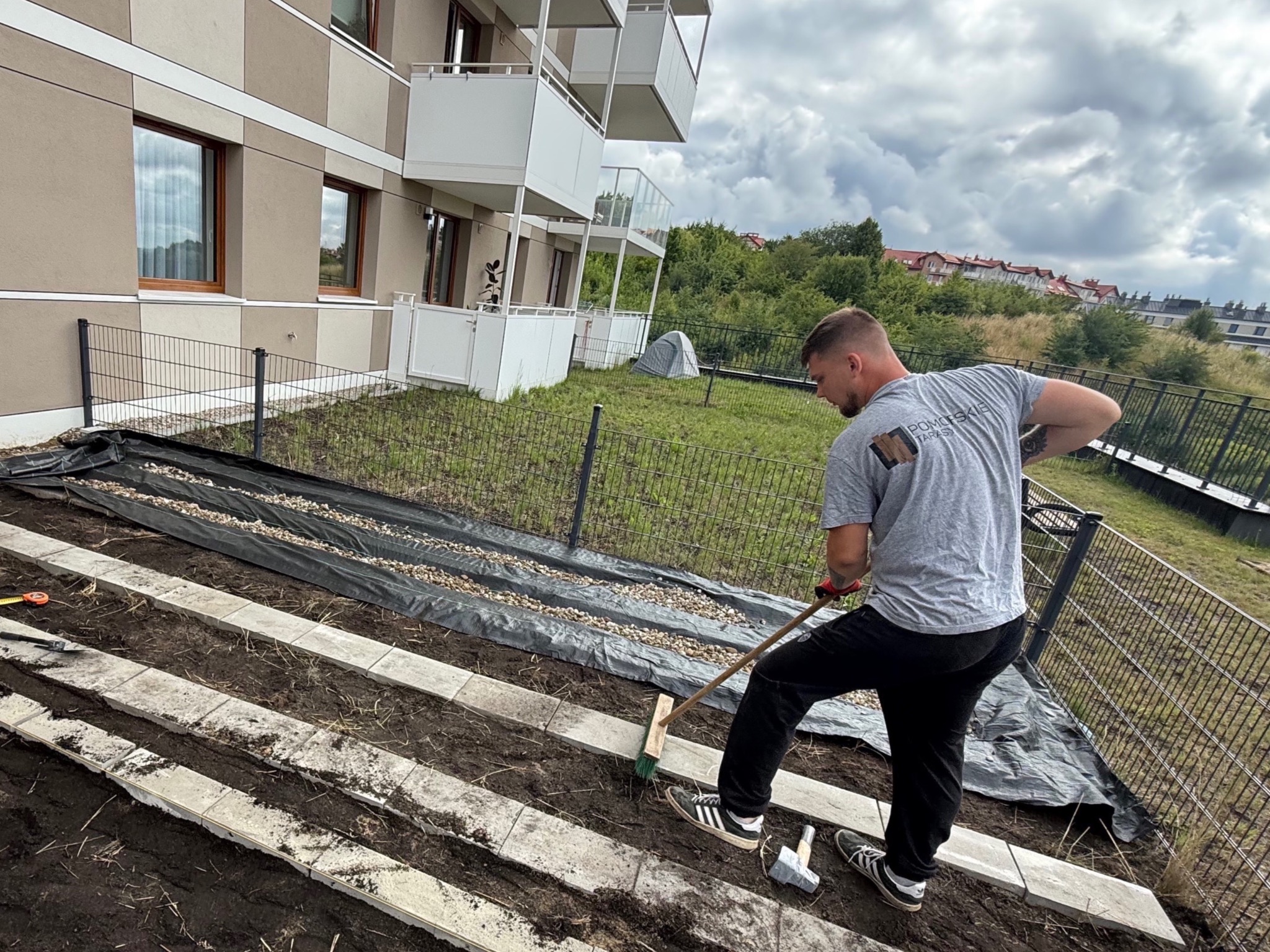 Pracownik wyrównuje podłoże pod taras z płyt betonowych. Widoczne narzędzia, geowłóknina i budynek mieszkalny w tle. Praca w toku.