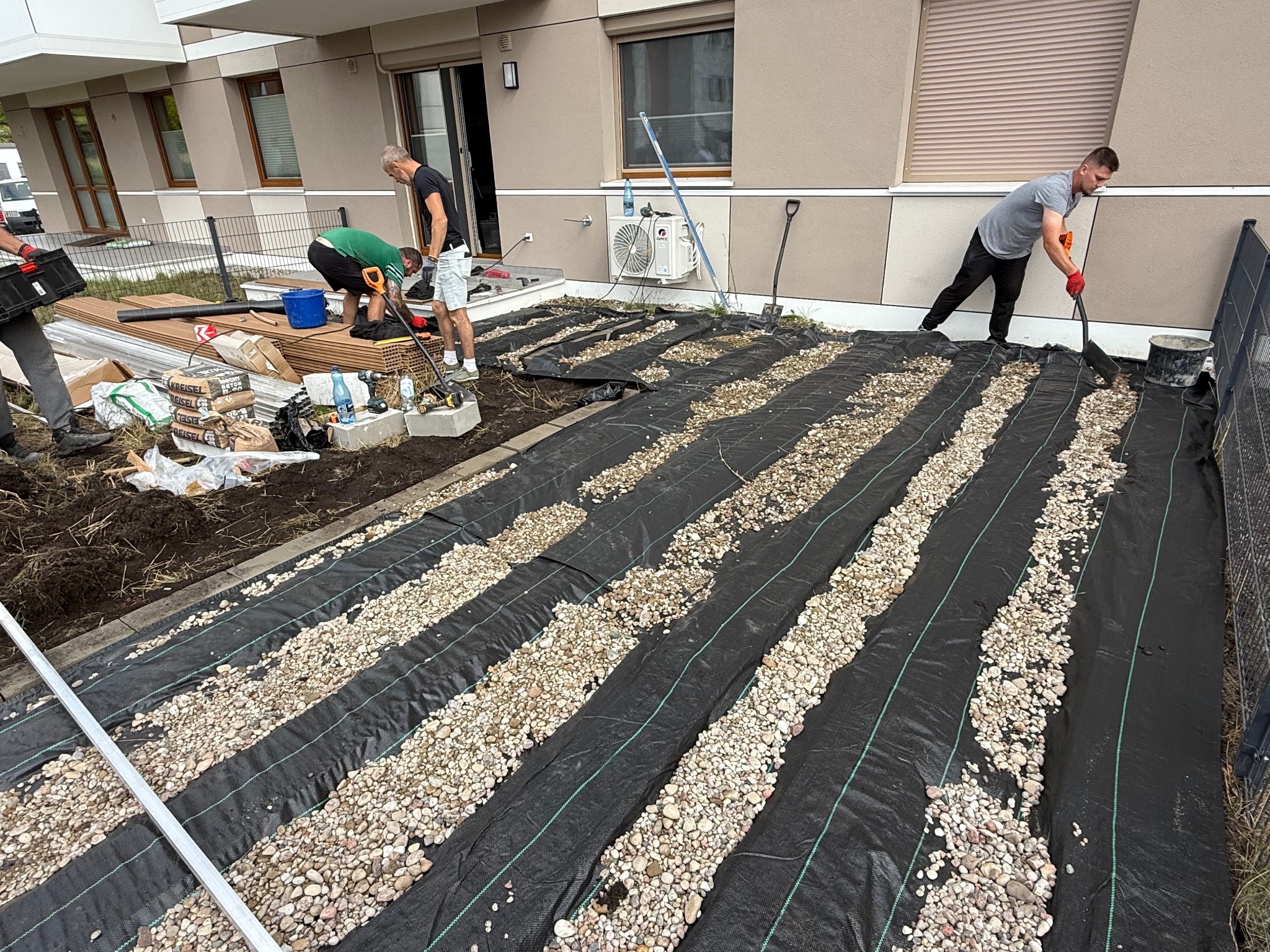 Montaż tarasu: robotnicy rozkładają geowłókninę i kamień drenażowy na przygotowanym podłożu, widoczne narzędzia i materiały budowlane, etap prac.