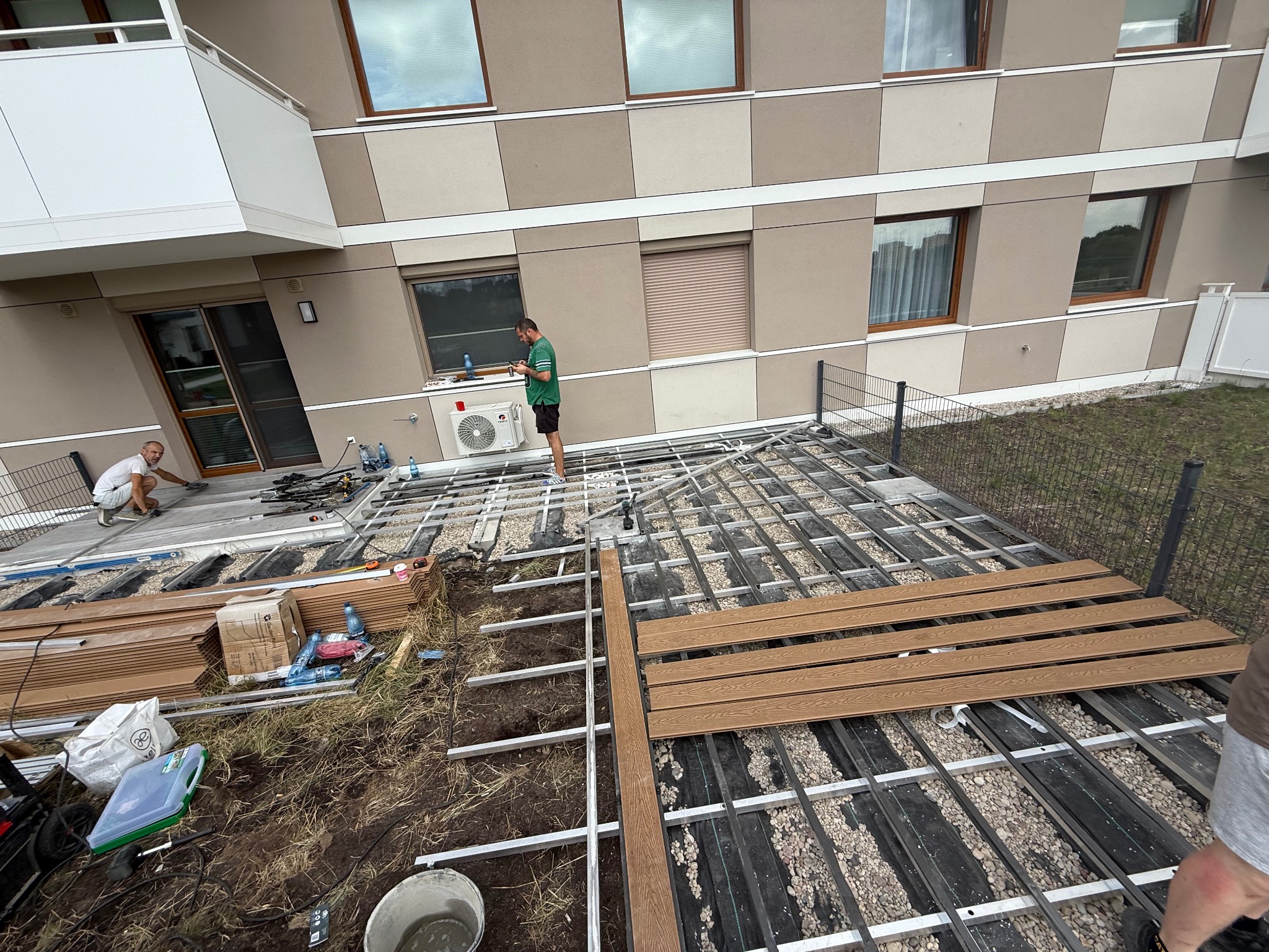 Montaż tarasu kompozytowego na balkonie budynku wielorodzinnego. Widoczne elementy konstrukcyjne i deski w kolorze drewna. Prace w toku, narzędzia i materiały na ziemi.