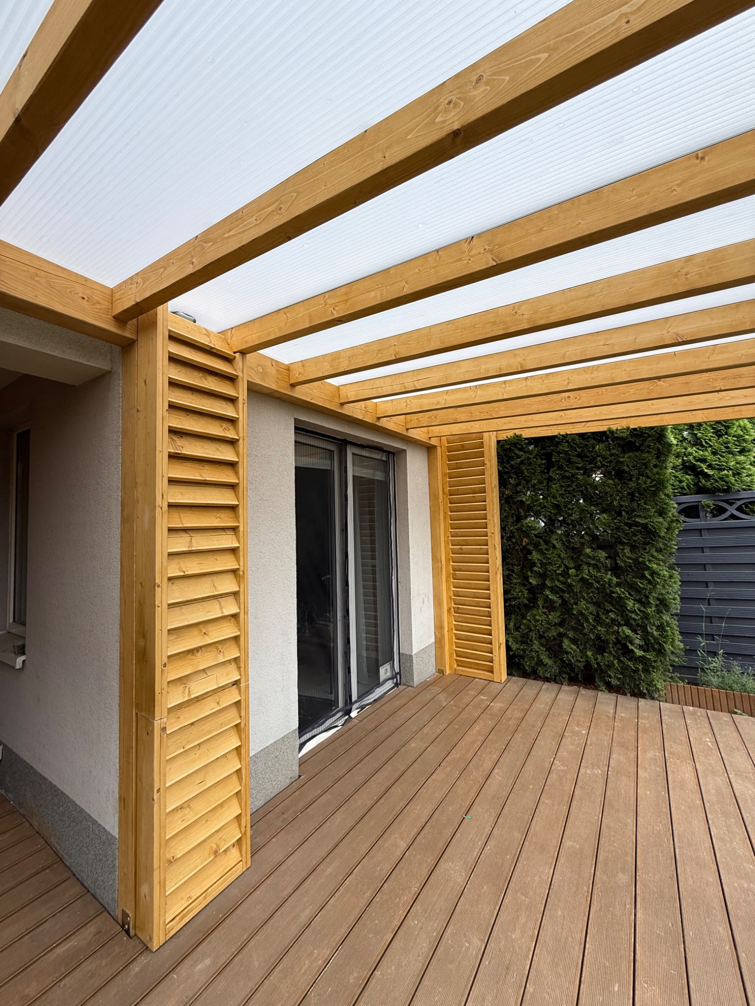 Drewniana pergola z zadaszeniem z poliwęglanu, połączona z budynkiem, z drewnianą podłogą tarasową. Realizacja z Gdańska.