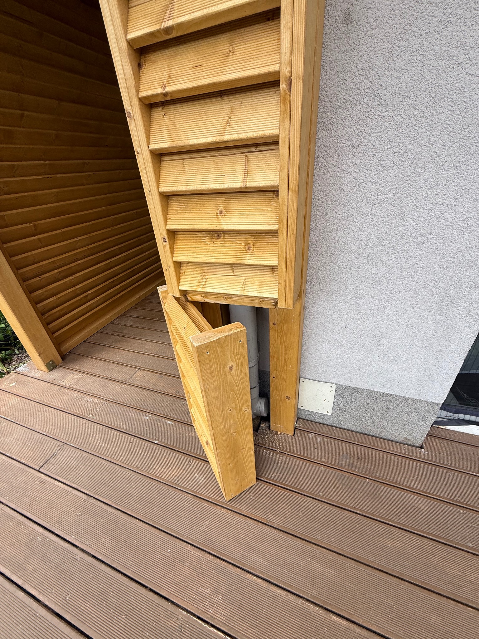 Drewniana zabudowa rury spustowej na tarasie kompozytowym, z otwieranym panelem dostępowym. Widoczna faktura desek tarasowych i fragment elewacji budynku.