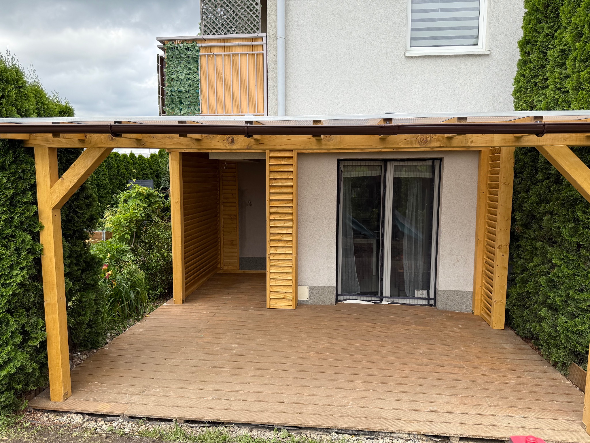 Drewniana pergola z zadaszeniem i ażurowymi ściankami, przylegająca do budynku z oknem i balkonem. Widok na taras z desek w otoczeniu zieleni.