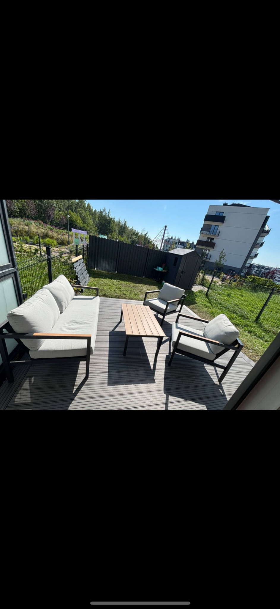 Nowoczesny taras z szarych desek kompozytowych. Zestaw mebli wypoczynkowych z jasnymi poduchami i stolikiem. W tle zielony trawnik i budynek mieszkalny.