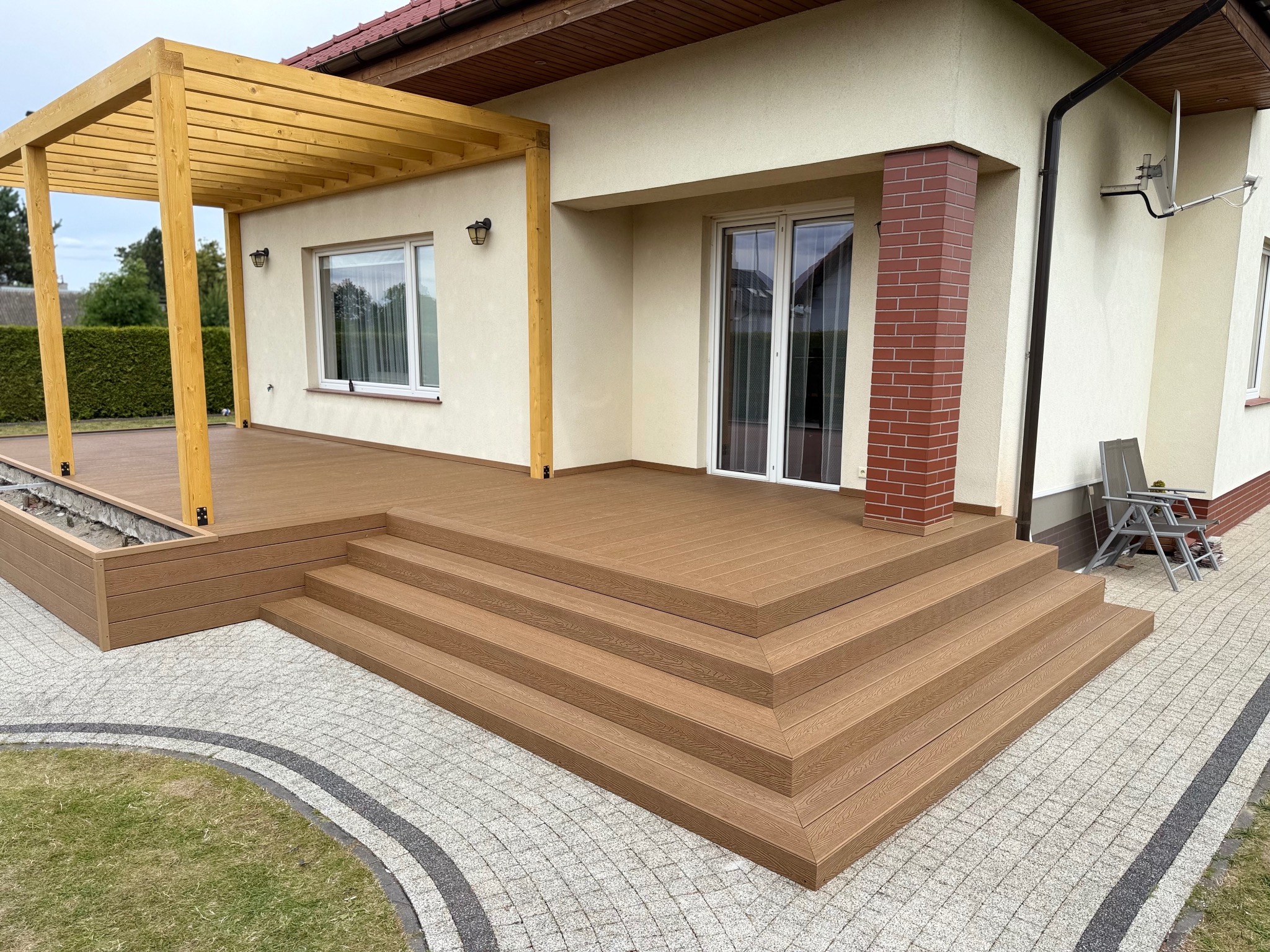 Beżowy taras kompozytowy z schodami i drewnianą pergolą przy domu z ceglanym filarem. Widok na kostkę brukową i zieleń w tle.