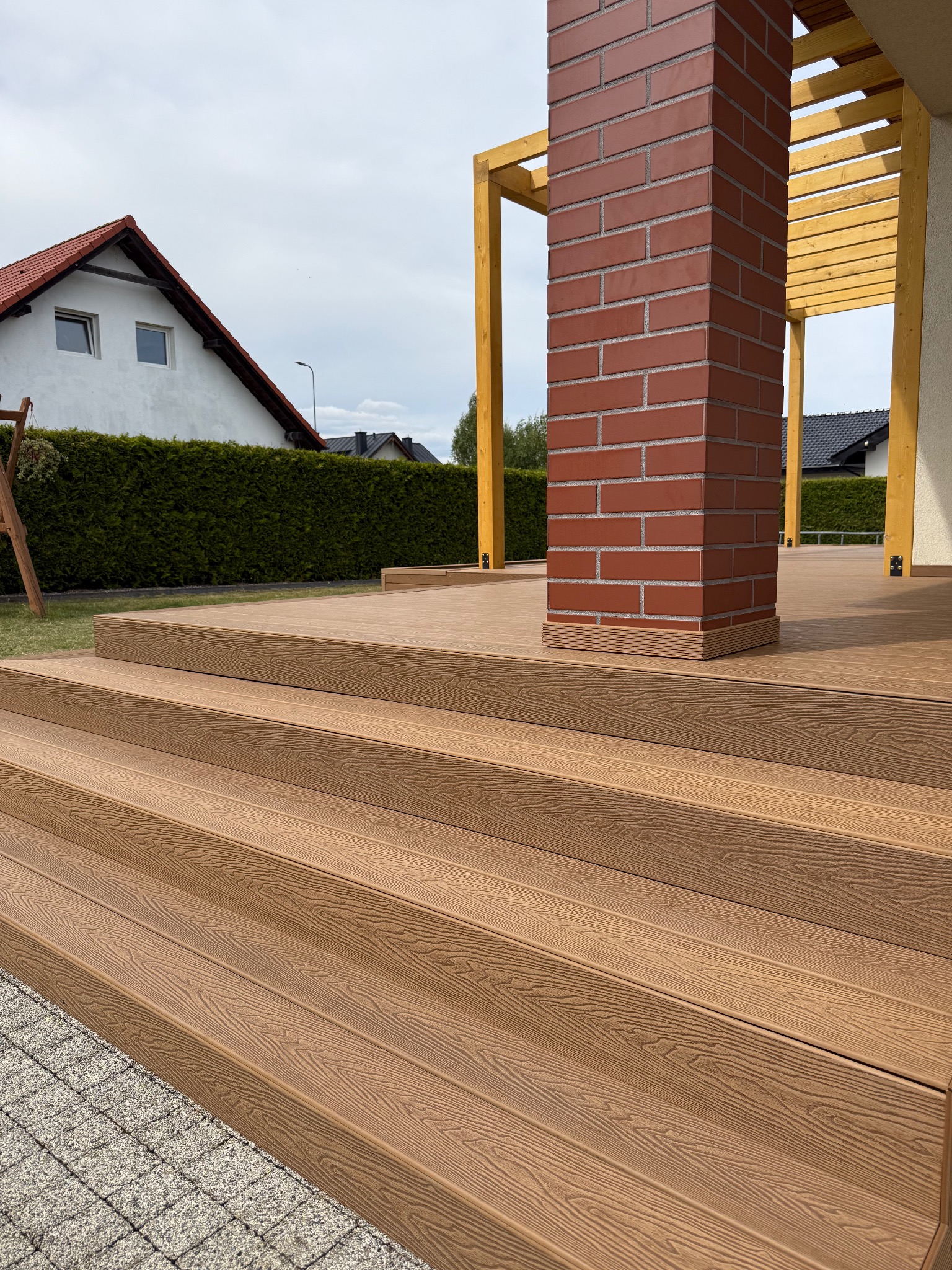 Schody z desek kompozytowych prowadzące na taras z ceglanym filarem i drewnianą konstrukcją. Widok na dom z ogrodem.