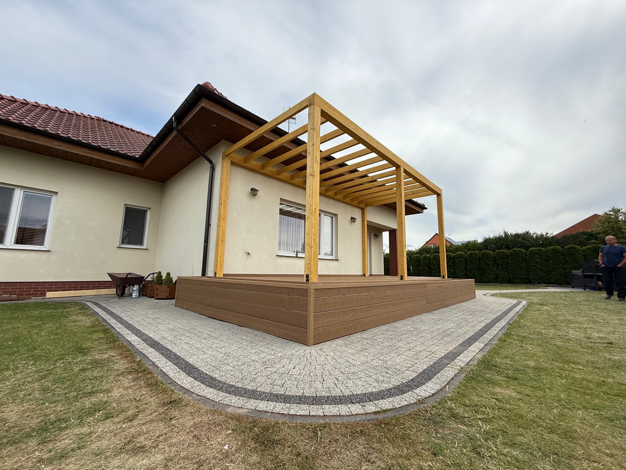 Nowoczesny taras kompozytowy z drewnianą pergolą, otoczony kostką brukową i trawnikiem. Widok na dom z jasną elewacją i czerwoną dachówką. Słoneczny dzień.