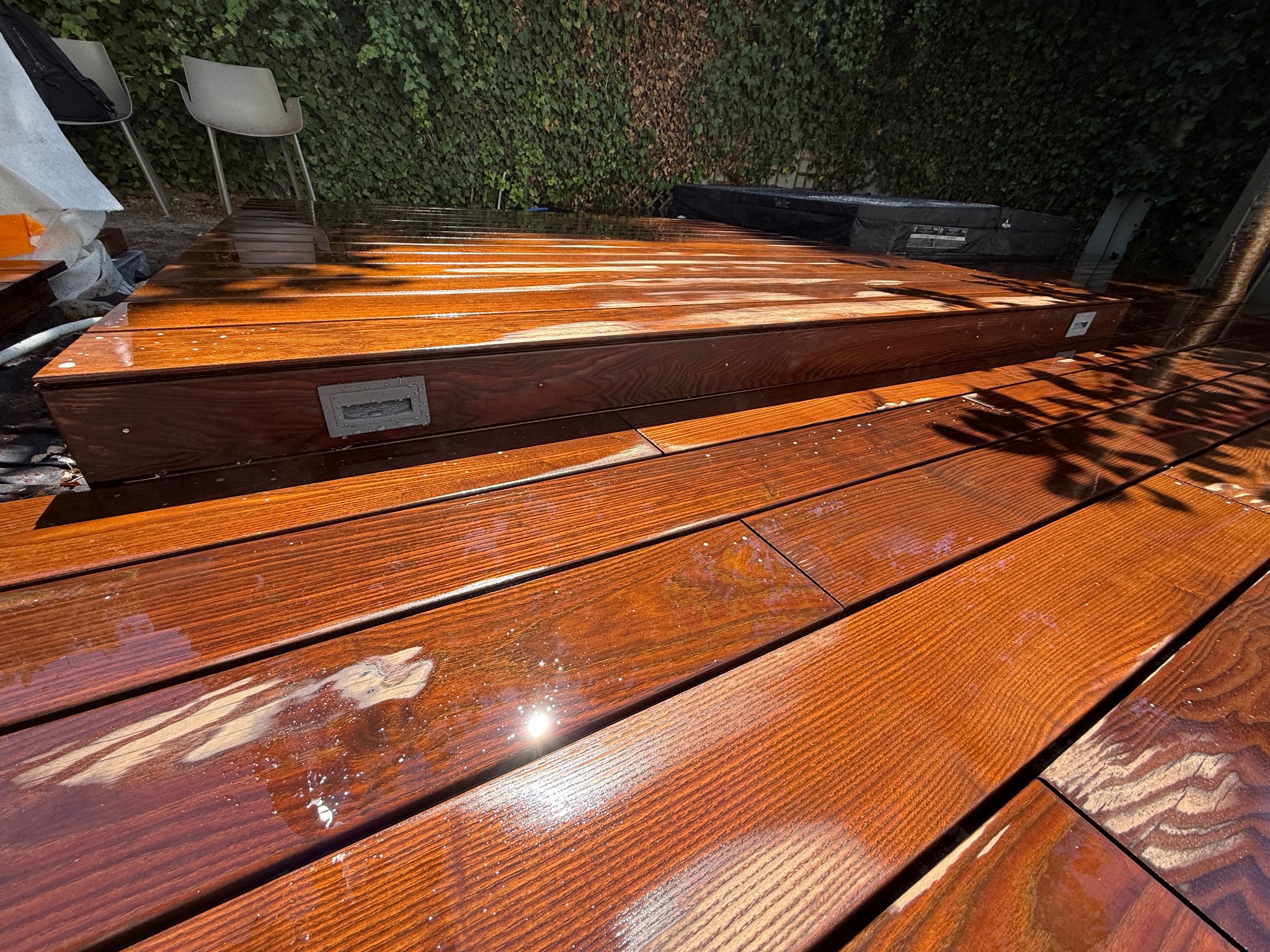 Błyszczący, świeżo polakierowany taras drewniany z oświetleniem podestowym i zielonym tłem z bluszczu. Widoczne ślady lakieru i odbicia światła.