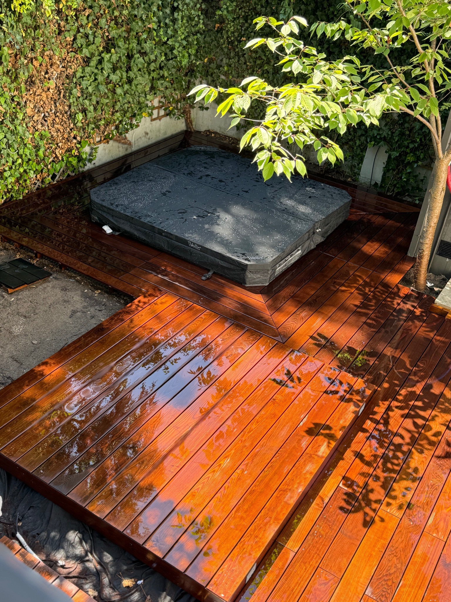 Błyszczący, drewniany taras z jacuzzi w ogrodzie, otoczony zielenią. Widoczne detale desek i odbicia światła. Ujęcie z góry ukazuje całą konstrukcję.