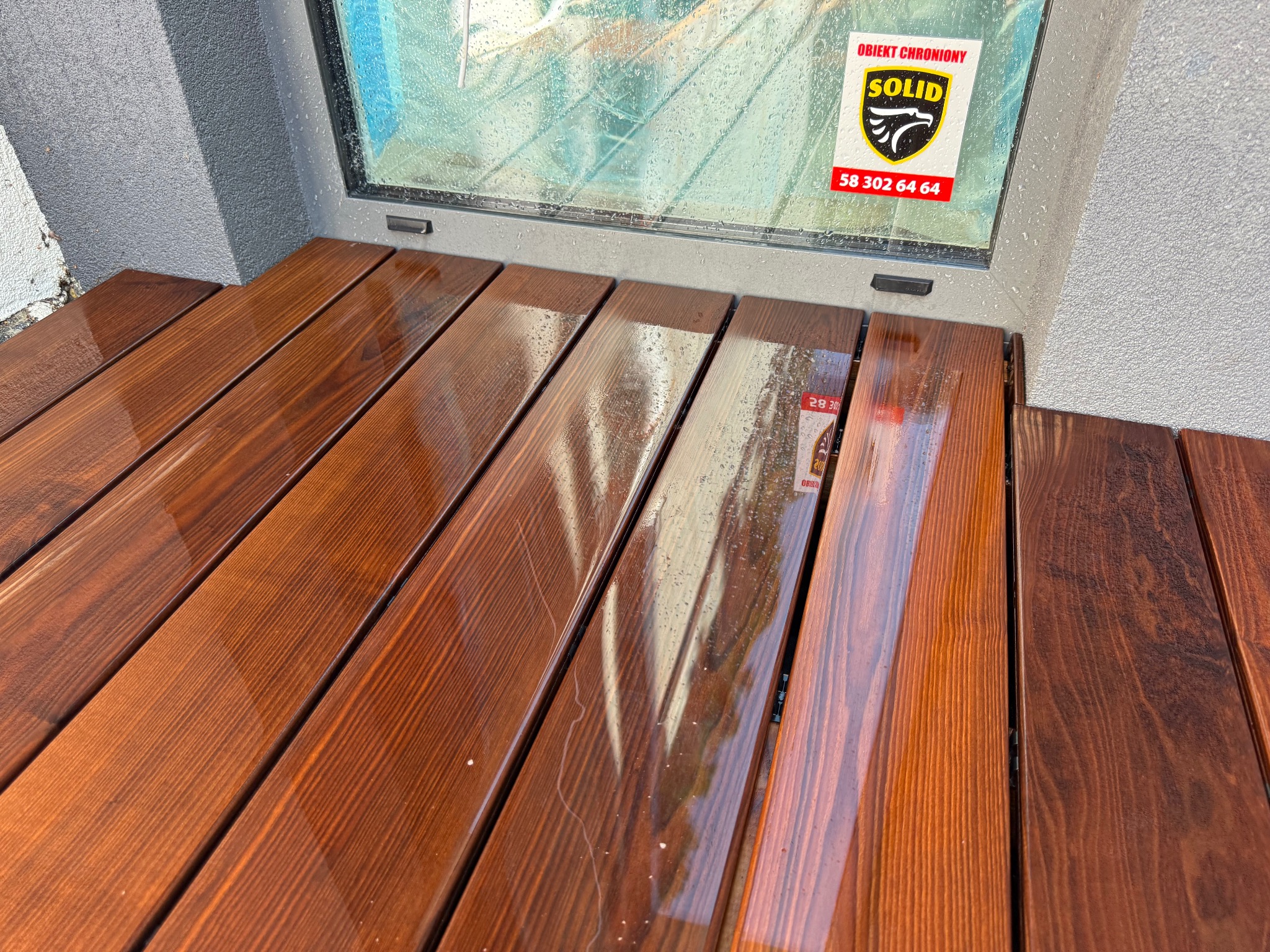 Mokry, drewniany taras z desek w odcieniach brązu, przylegający do okna z naklejką 'Obiekt Chroniony SOLID'. Widoczne krople deszczu na powierzchni drewna i szybie.