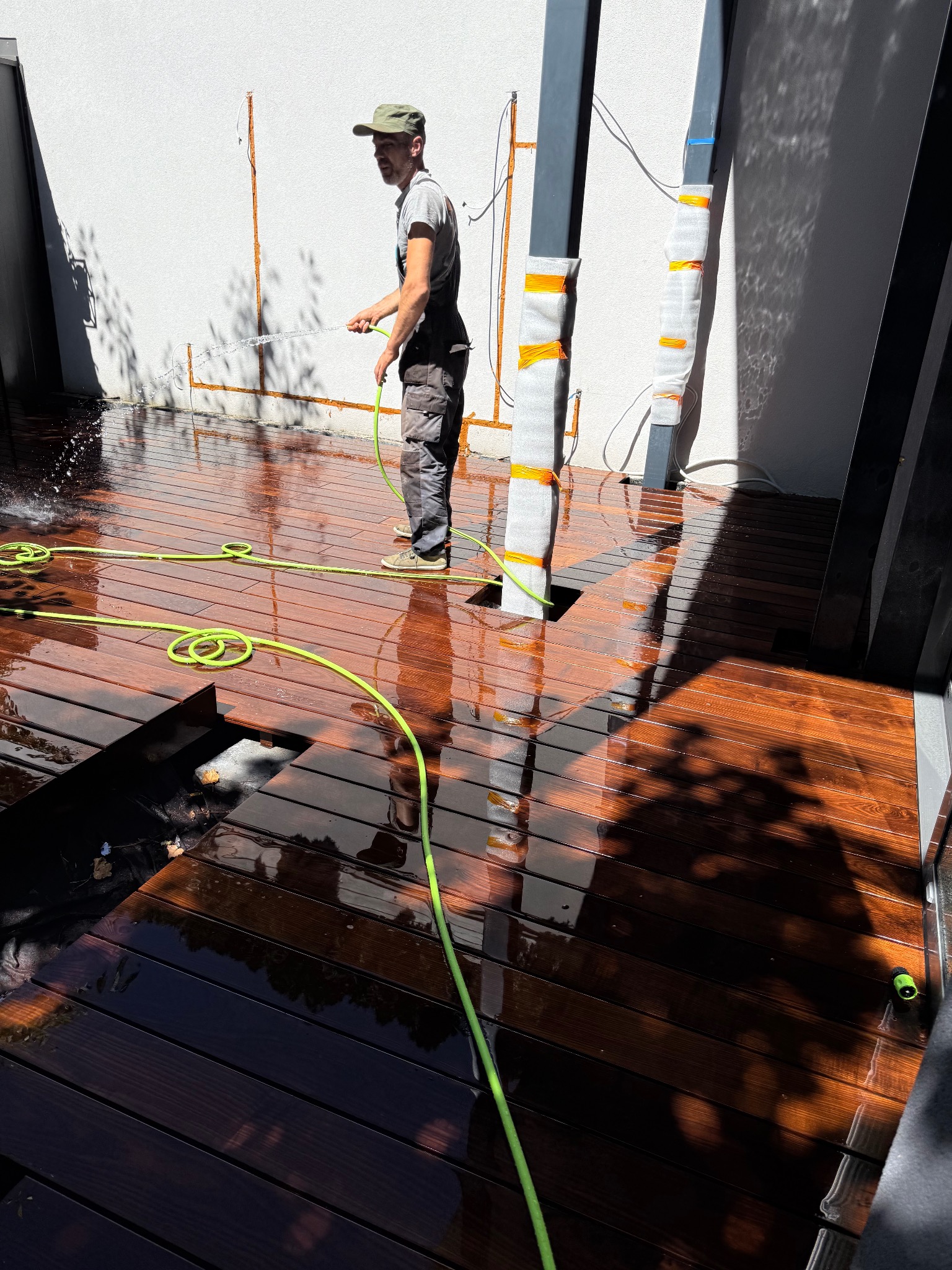 Czyszczenie tarasu z drewna egzotycznego z użyciem węża ogrodowego. Widoczne elementy konstrukcji pergoli i fragment niezabudowanej przestrzeni pod tarasem.