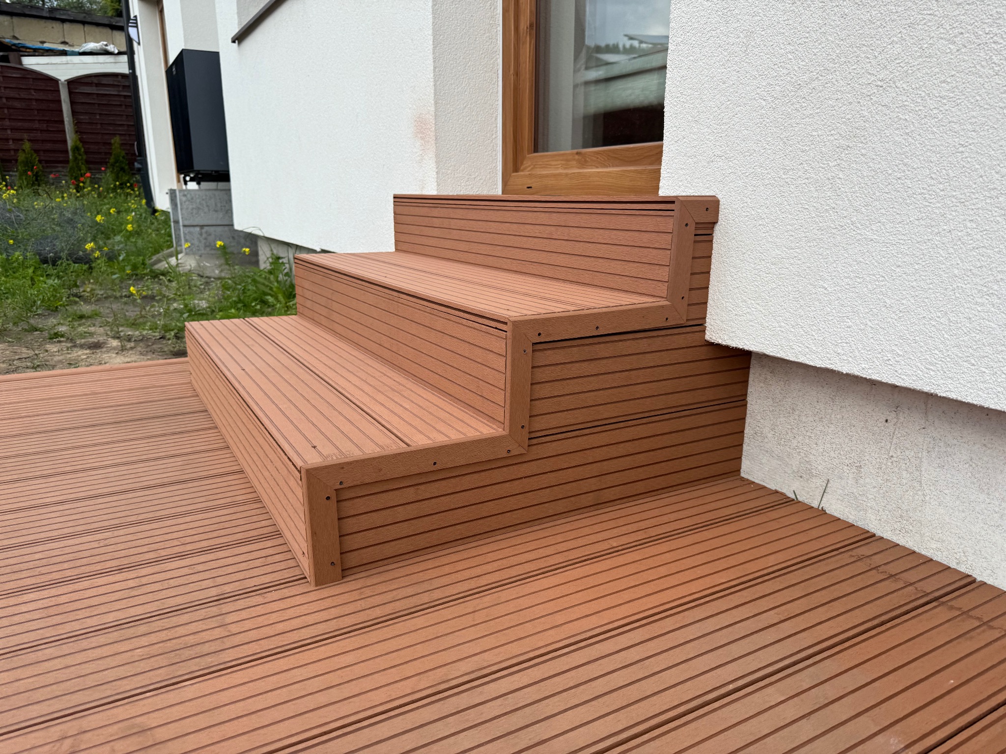 Schody i taras z deski kompozytowej w kolorze brązowym, przylegające do białego budynku z oknem. Widoczne mocowania desek.