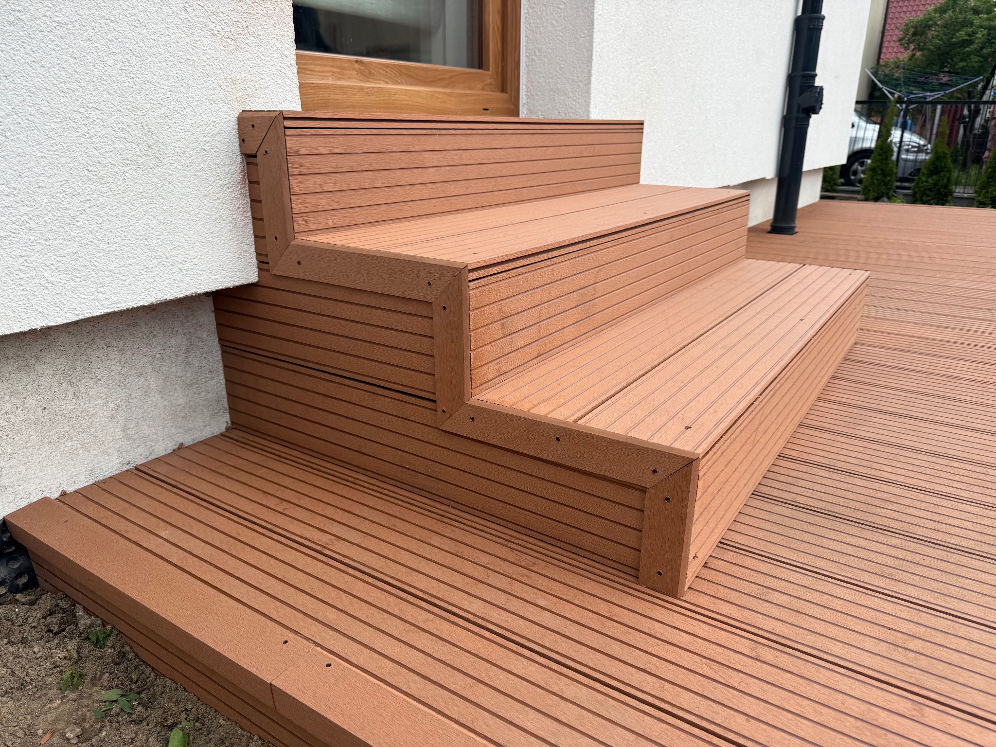 Brązowe schody z desek kompozytowych prowadzą na taras, przylegając do białej ściany budynku. Widoczne detale łączeń i faktura desek. Ujęcie z dołu.