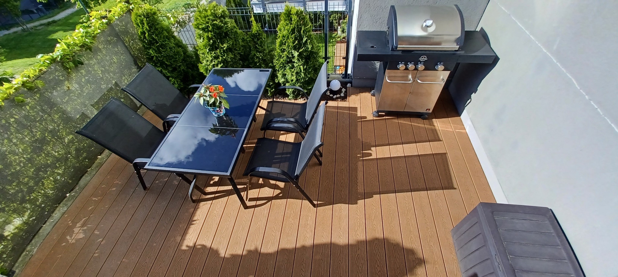 Jasny taras kompozytowy z meblami ogrodowymi i grillem. Widok z góry na brązowe deski, czarne krzesła i odbicie nieba w blacie stołu.
