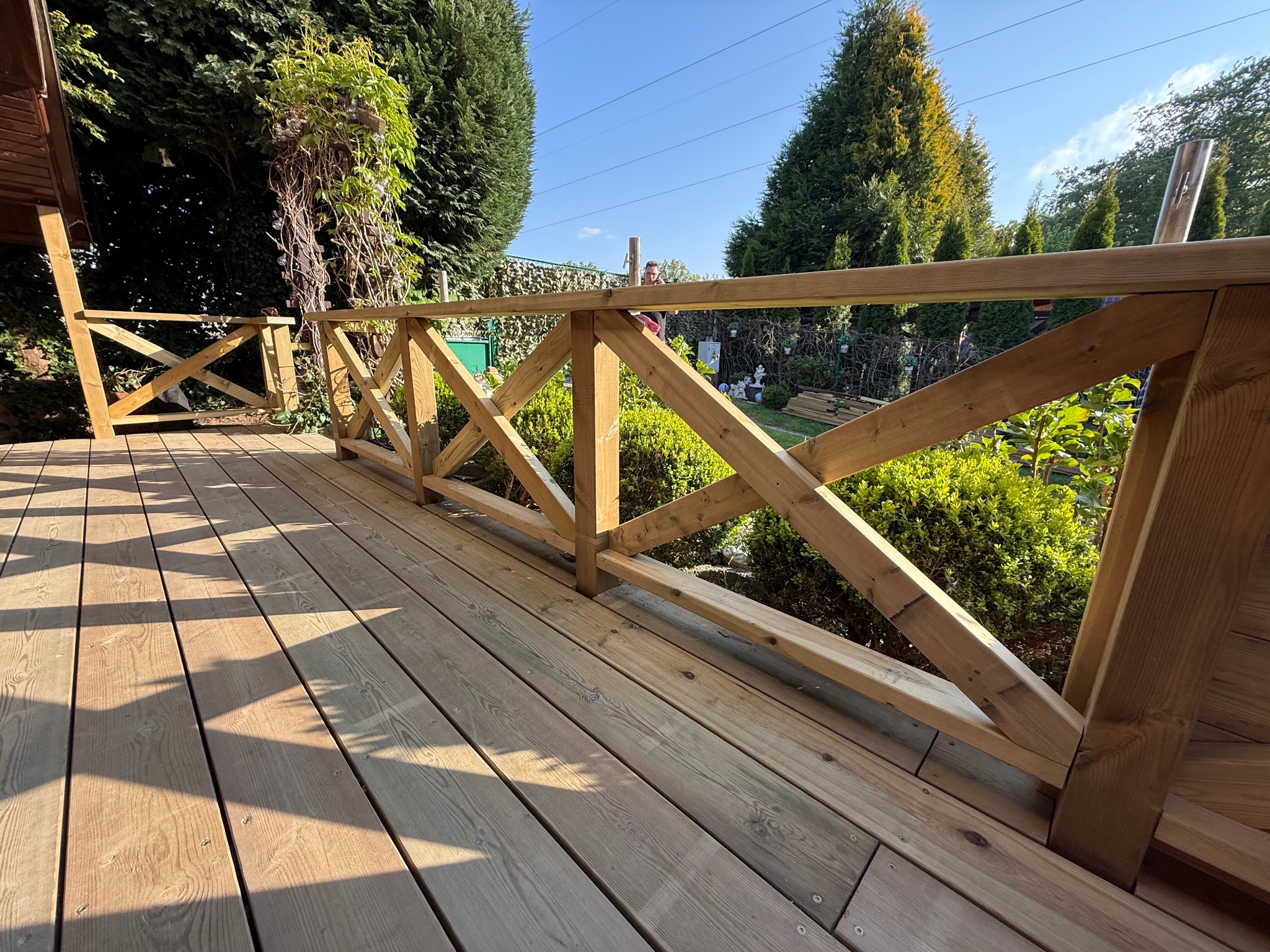 Jasny taras z drewnianą balustradą w kształcie krzyży, otoczony zielenią i drzewami. Widok z poziomu podłogi, słoneczny dzień.