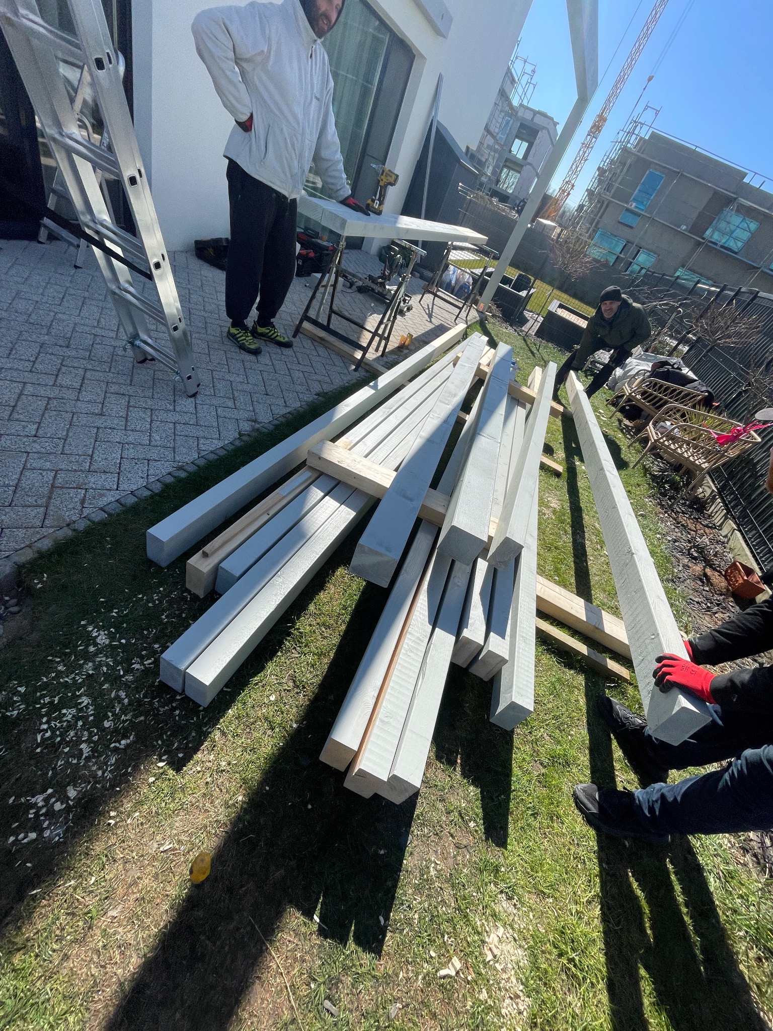 Przygotowanie elementów konstrukcyjnych na trawie do montażu, prawdopodobnie pergoli lub altany, z pracownikami w tle i drabiną obok.