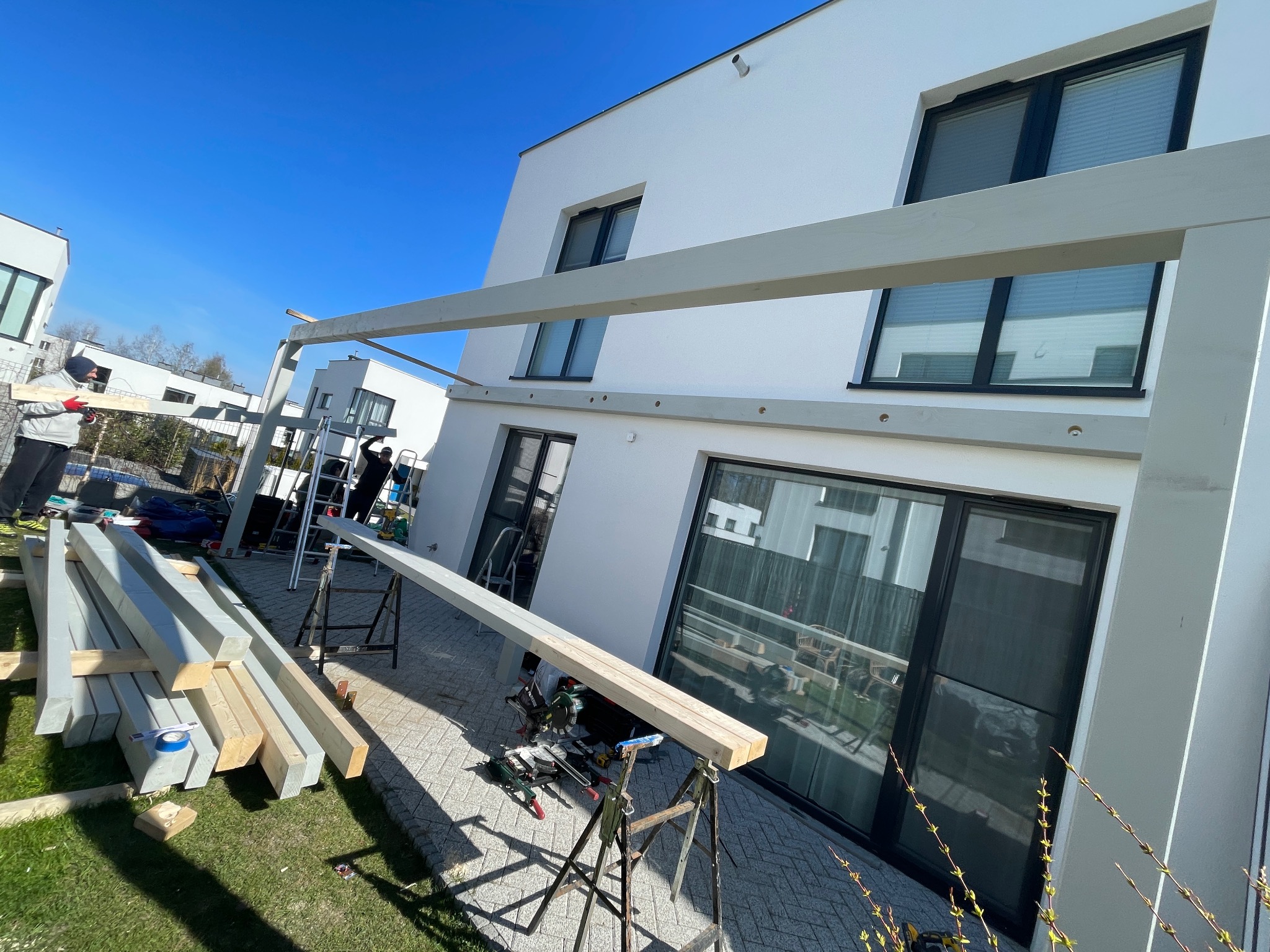 Montaż drewnianej pergoli przy nowoczesnym domu. Widoczne narzędzia, przygotowane elementy konstrukcji i pracownicy w trakcie pracy. Słoneczny dzień.