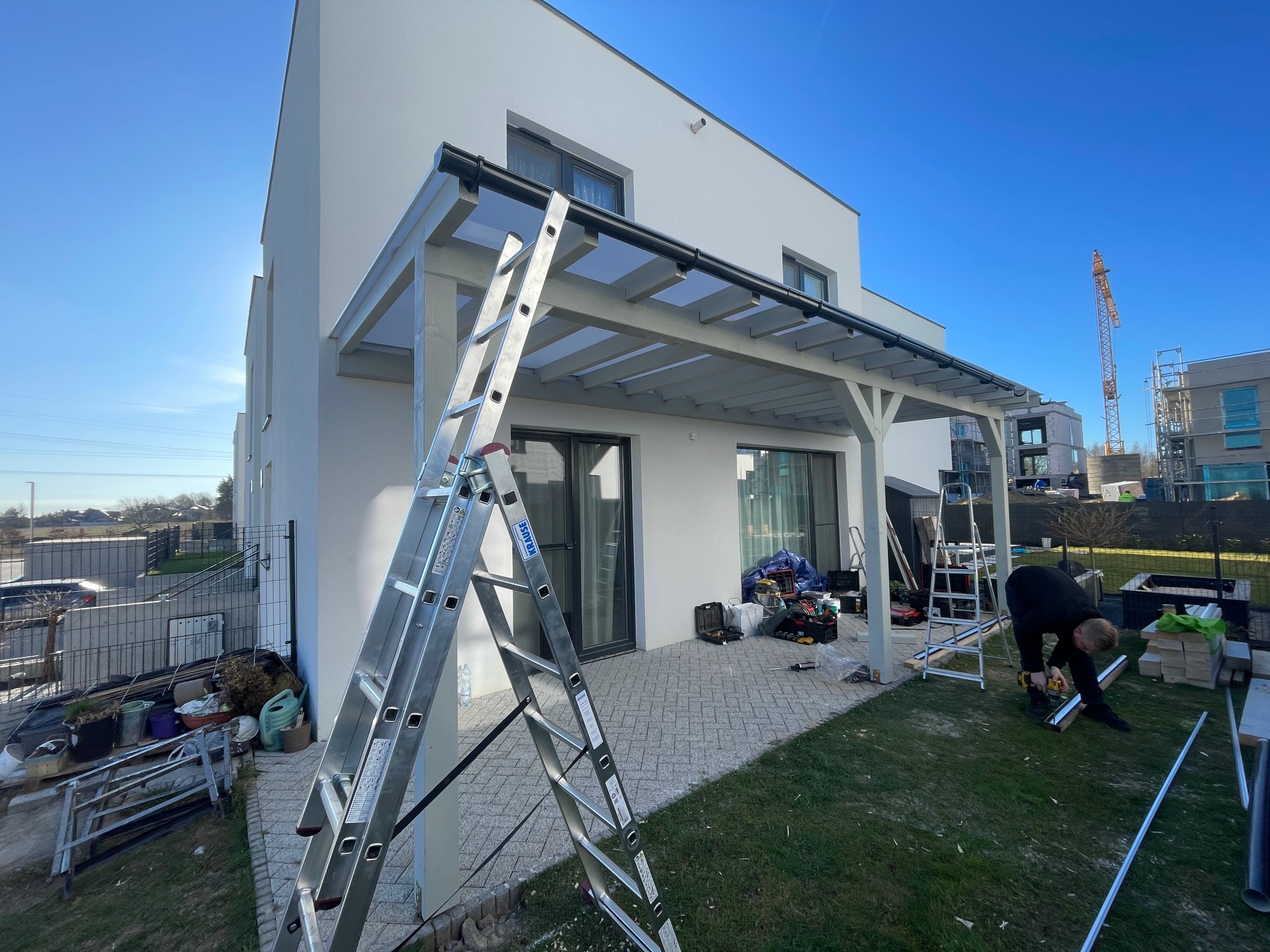 Montaż pergoli przy nowoczesnym domu. Widoczna drabina, narzędzia i pracownik przycinający element konstrukcyjny na trawniku w słoneczny dzień. Żuraw budowlany w tle.