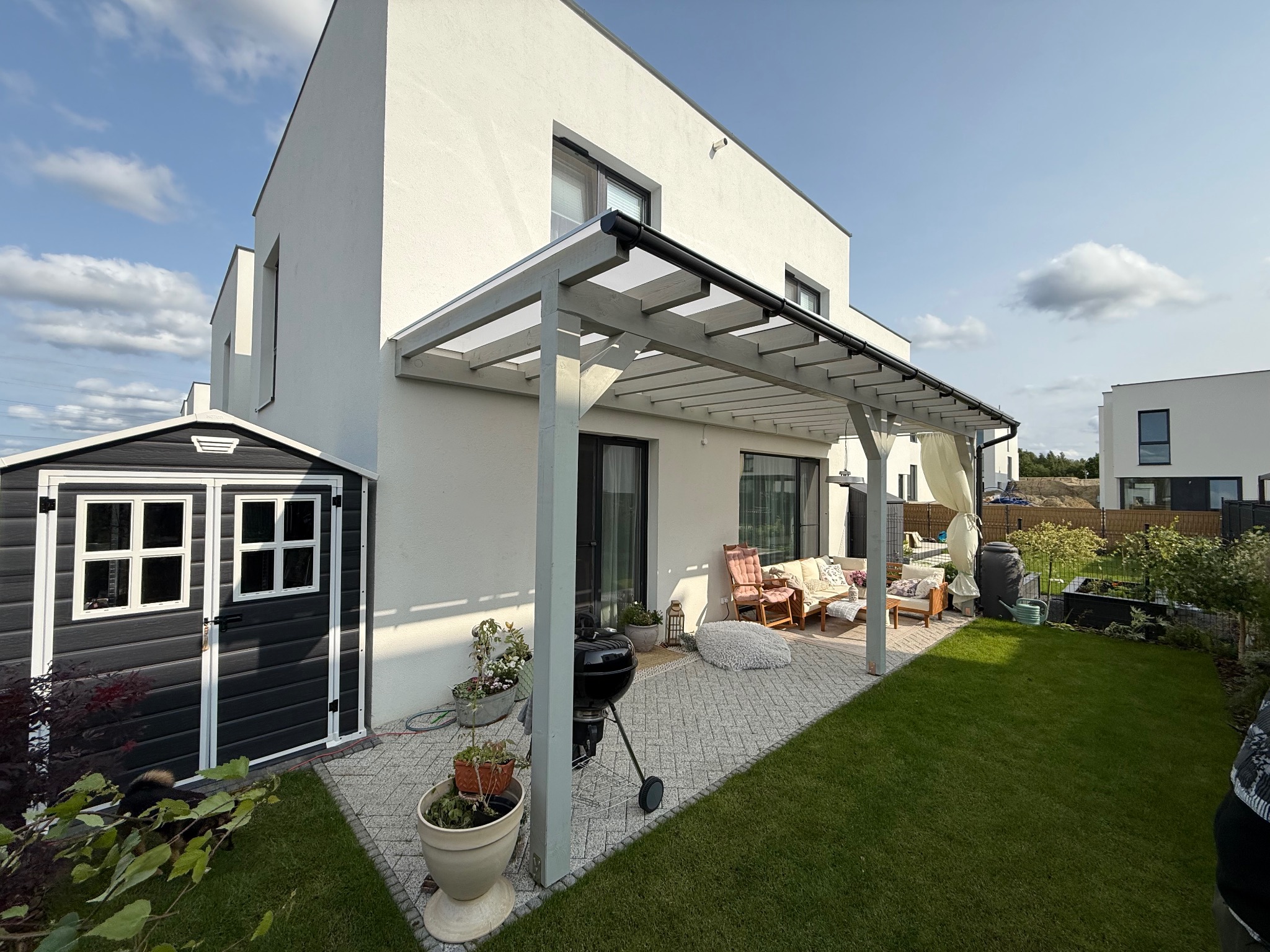 Nowoczesna pergola z przezroczystym zadaszeniem przy białym domu. Umeblowany taras z grillem i widokiem na zadbany trawnik. Obok stoi szara drewutnia.
