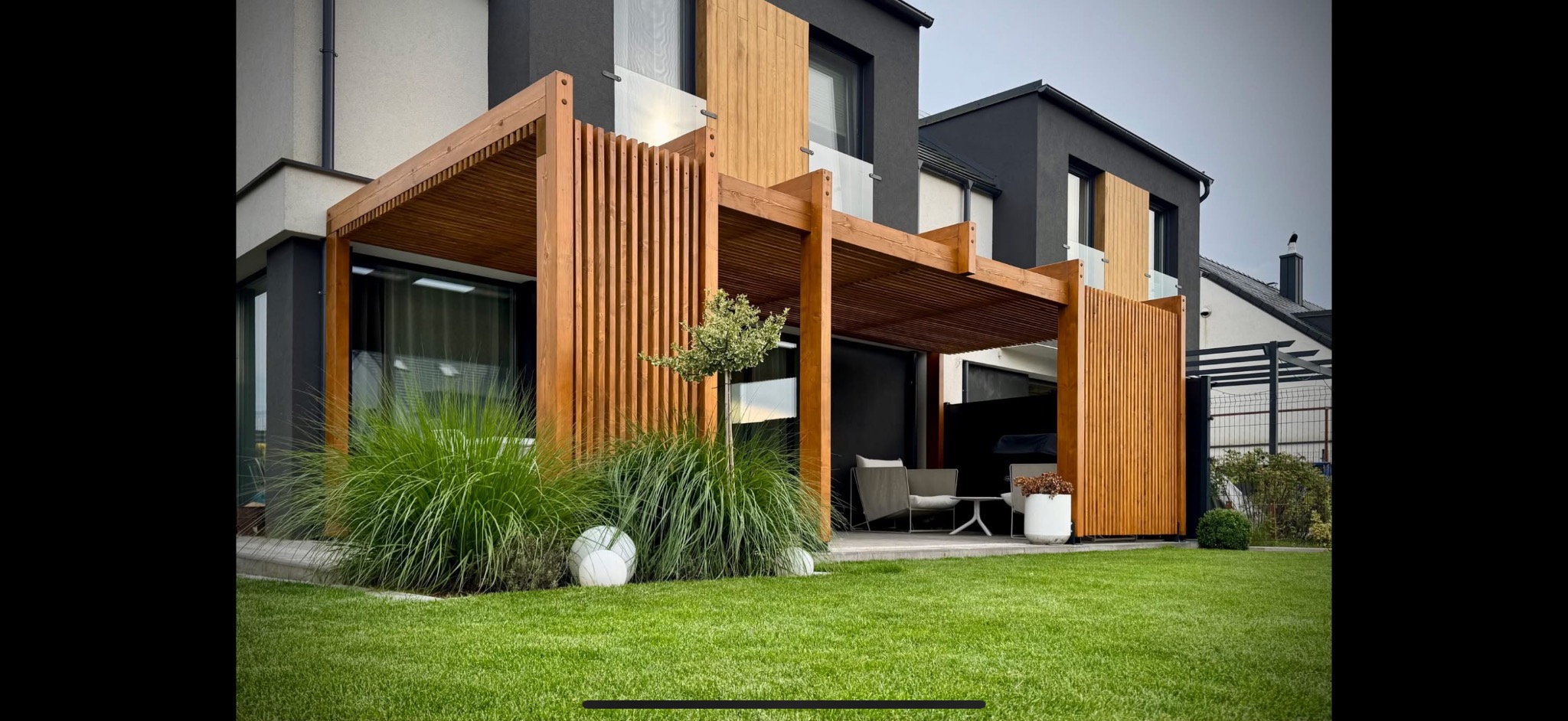 Drewniana pergola z pionowymi listwami, przylegająca do nowoczesnego domu z czarną elewacją. Na trawniku dekoracyjne trawy i kule. Widoczne meble ogrodowe.