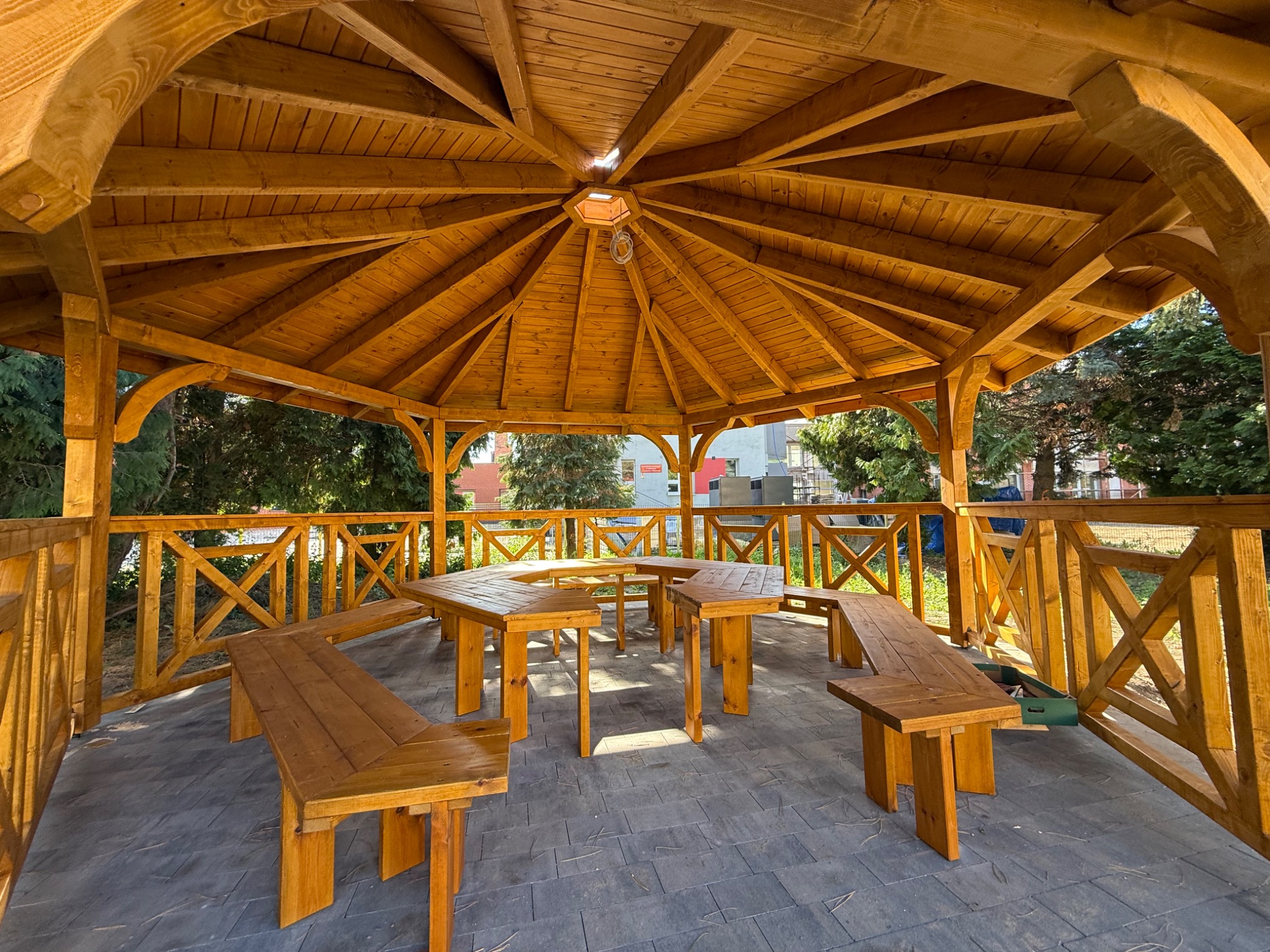 Drewniana altana z meblami ogrodowymi wewnątrz, widok z zewnątrz. Konstrukcja z belek, zadaszenie wielospadowe, ażurowe balustrady. Kamienna posadzka.