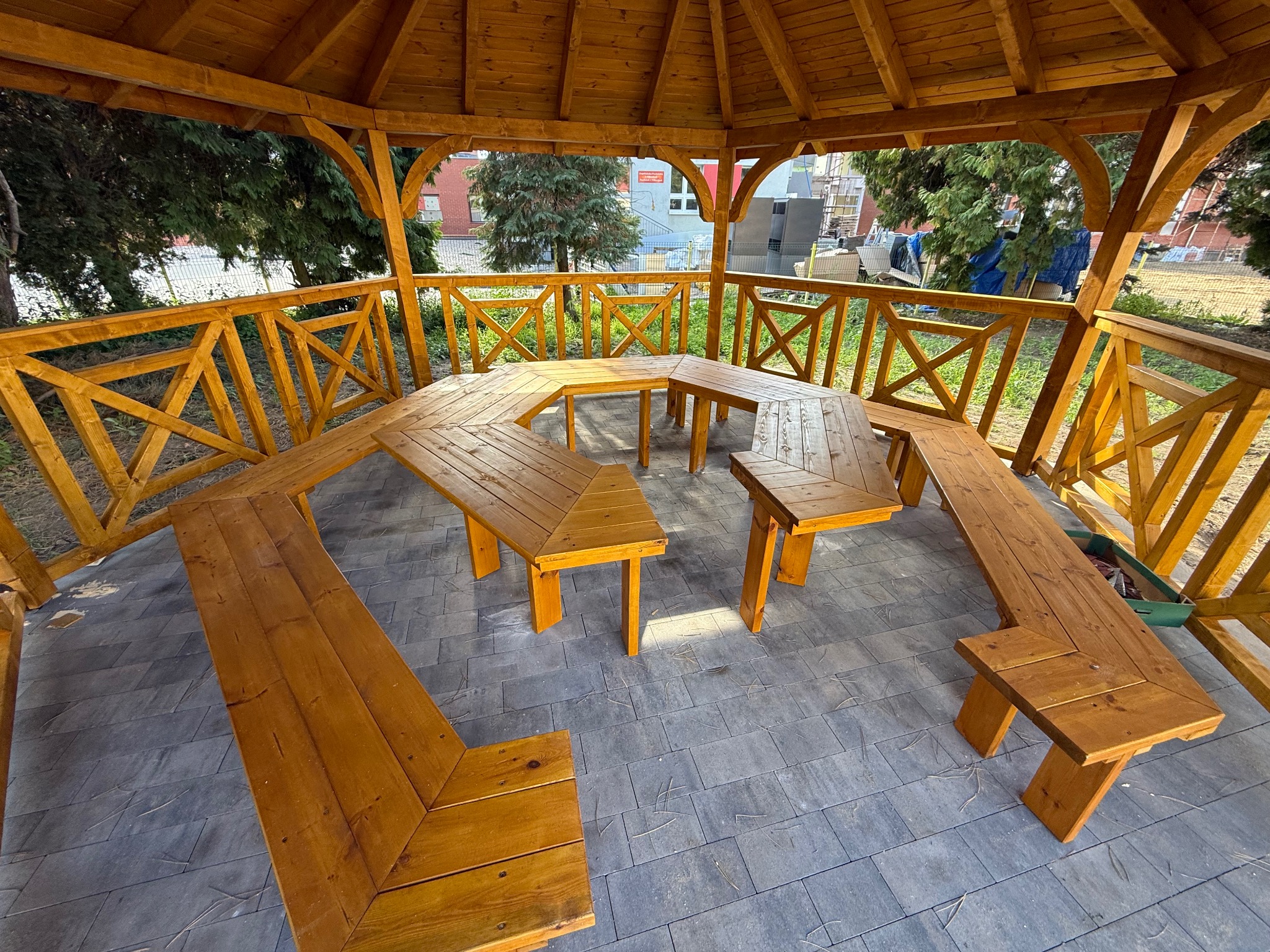 Drewniana altana z ławkami i stołem wewnątrz, widok z góry. Konstrukcja z belek, podłoga z szarej kostki. Na zewnątrz zieleń drzew i budynek w tle.