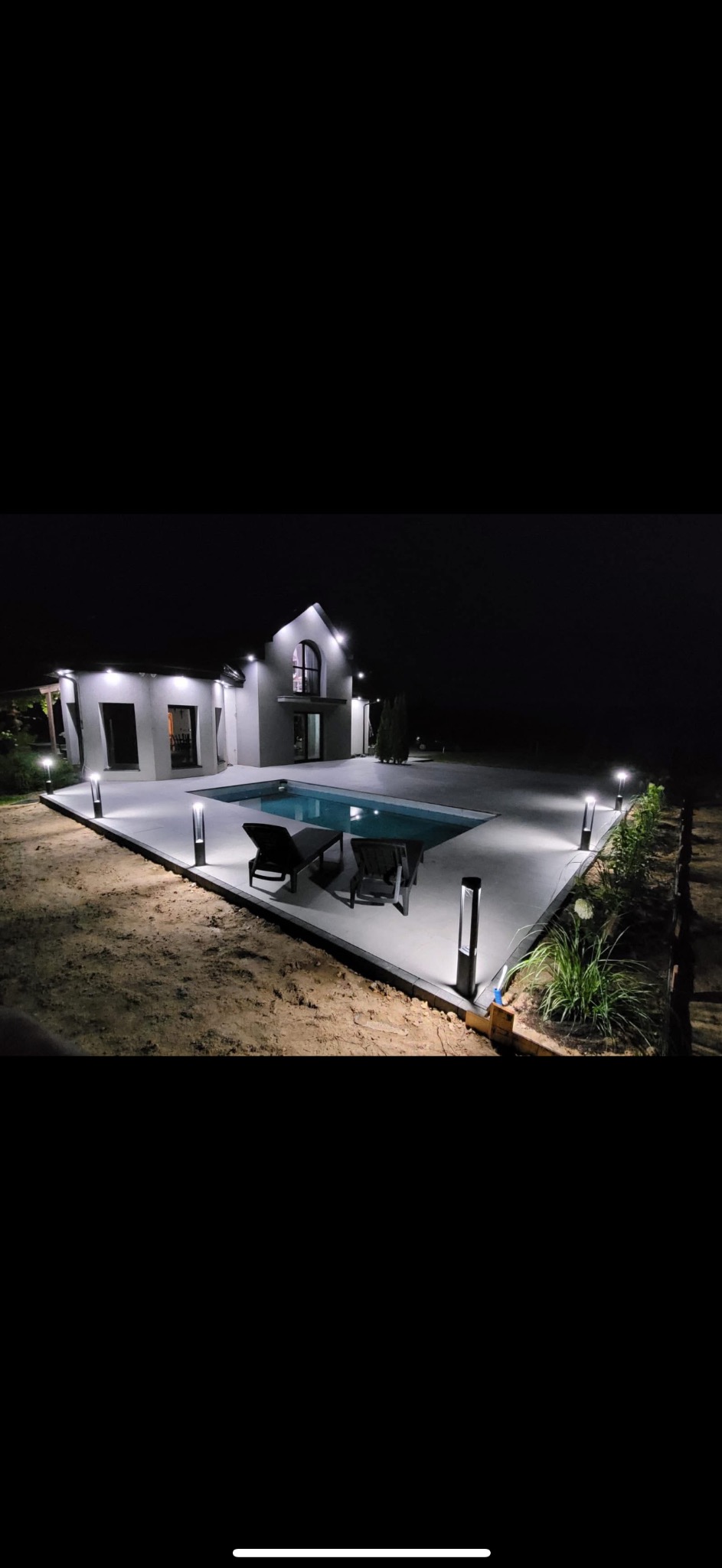 Nowoczesny dom z basenem oświetlonym nocą. Dwa leżaki przy basenie. Oświetlenie ścieżki lampami. Minimalistyczny design domu z dużymi oknami i oświetleniem zewnętrznym.